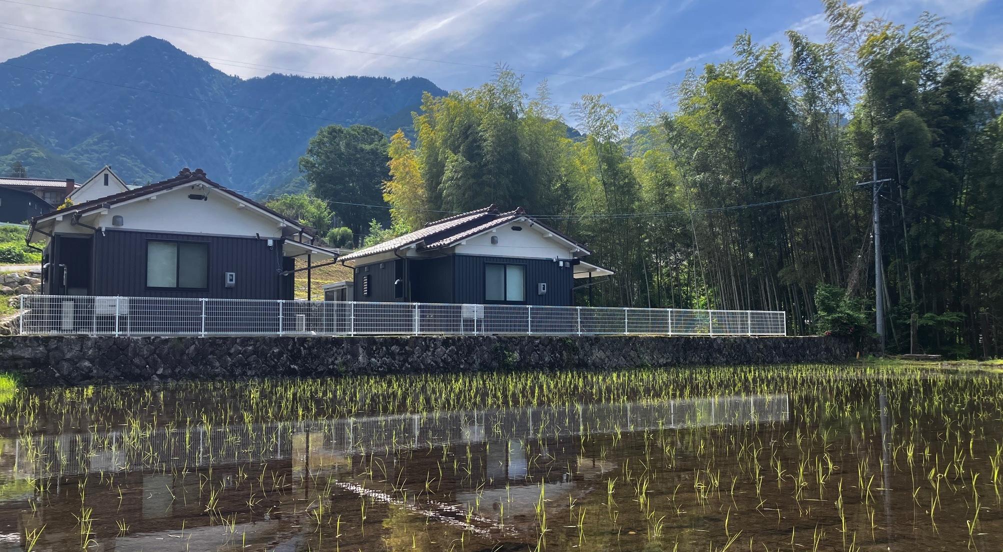 手前に水田が広がり水面に家と山が映る緑の山あいに建つ二軒の施設とその背後の竹林