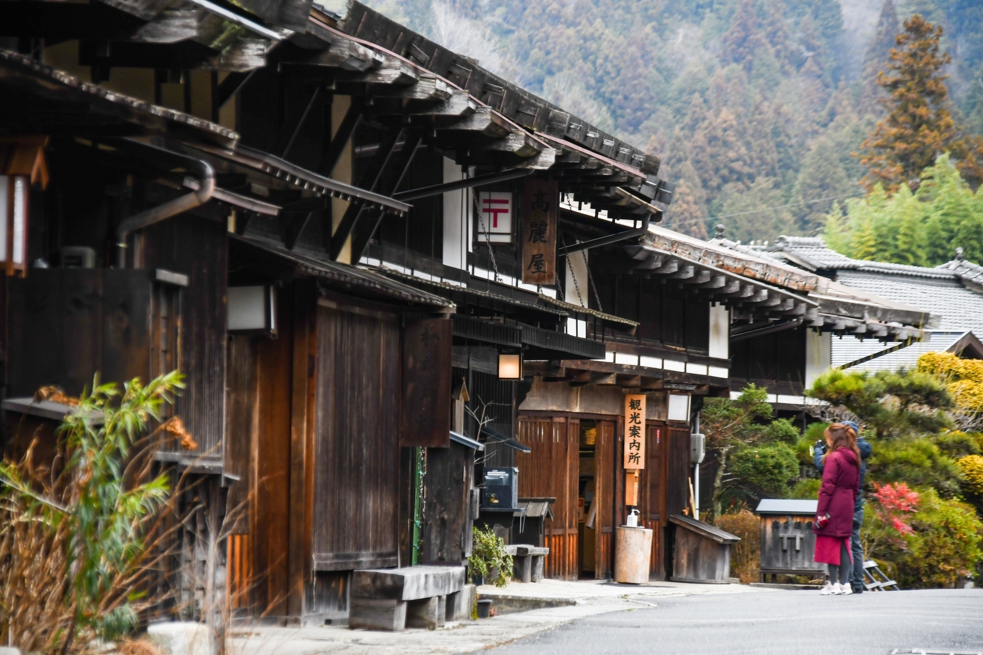 山を背景に古い木造の町家が並ぶ通りの前でコートを着た人物が歩いている場面