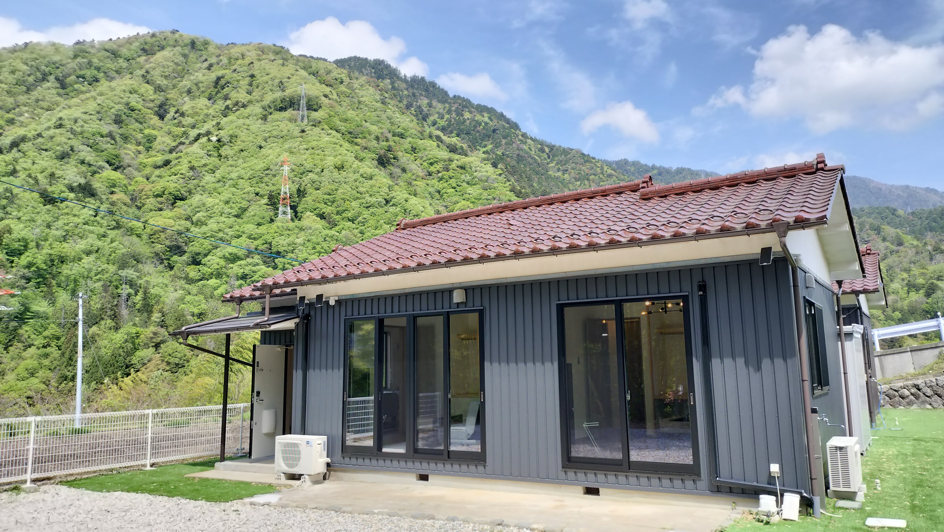 赤い瓦屋根とガラス引き戸の平屋が緑の山を背にして立つ家