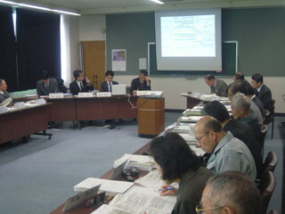 会議室で円形のテーブルに沿って座り資料に目を通しながらプロジェクターのスクリーンを前に議論する複数の参加者の様子