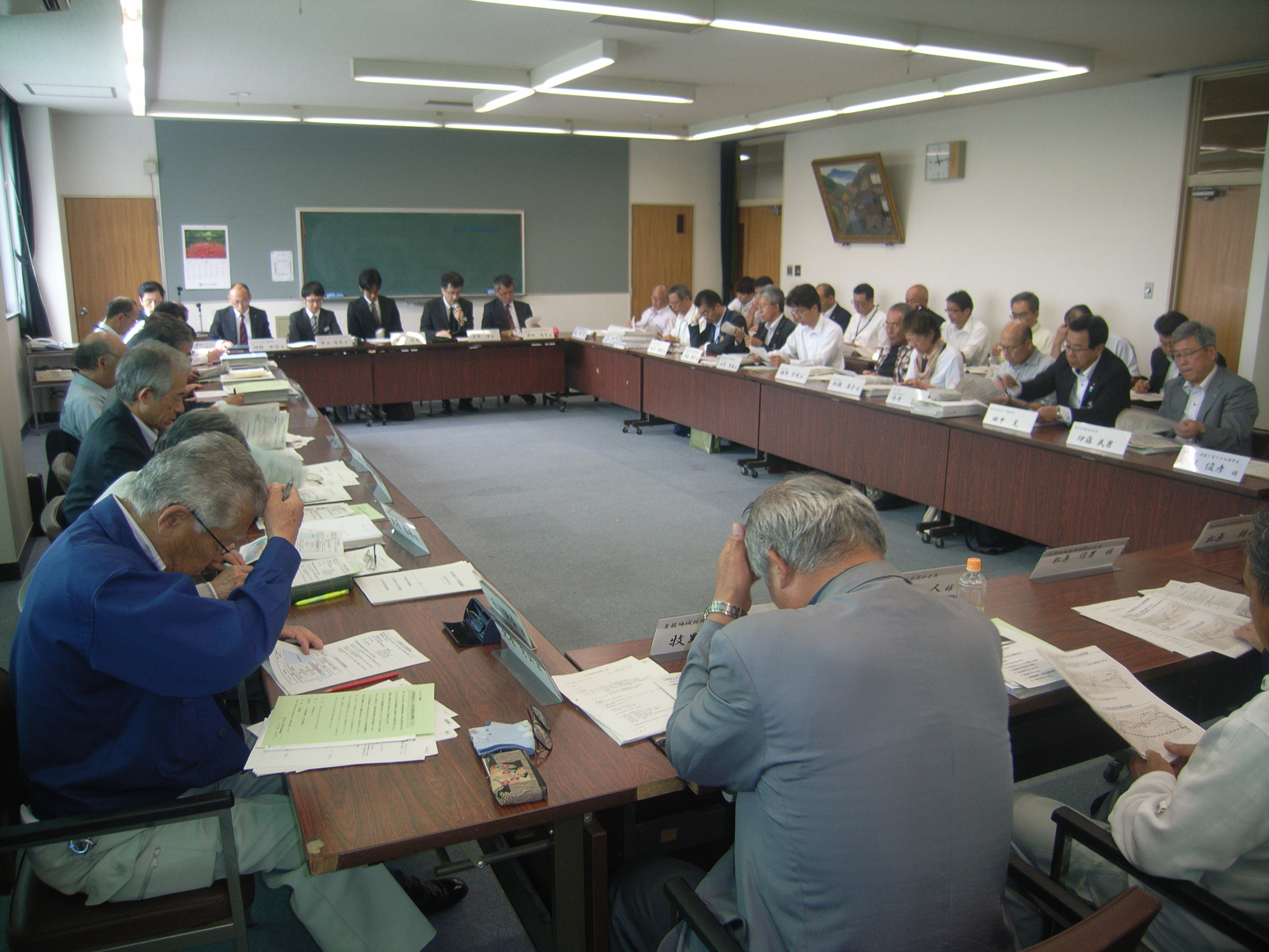 会議室で長い机を囲み資料を見ながら話し合う多数の参加者が座っている風景