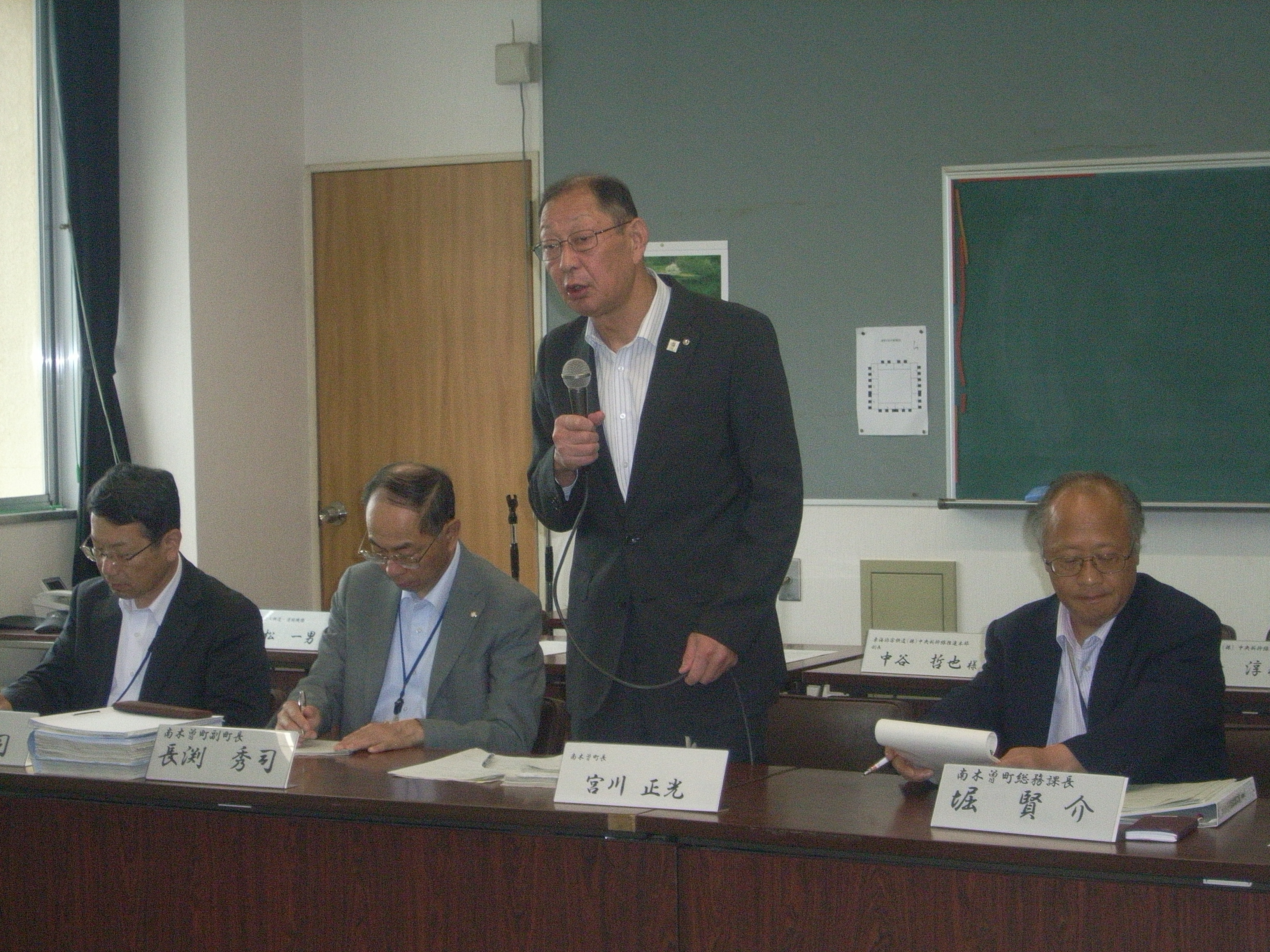 会議室の机に並んで座る数人の男性のうち中央の人物がマイクを持って立って発言している風景