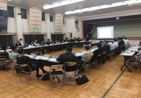 体育館のような広い室内で長方形に並べた机の周りに多くの参加者が座りプロジェクターで資料を投影している会議の様子