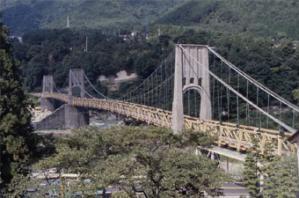 緑に囲まれた渓谷をまたぐ古い吊り橋とその周辺を見下ろす風景