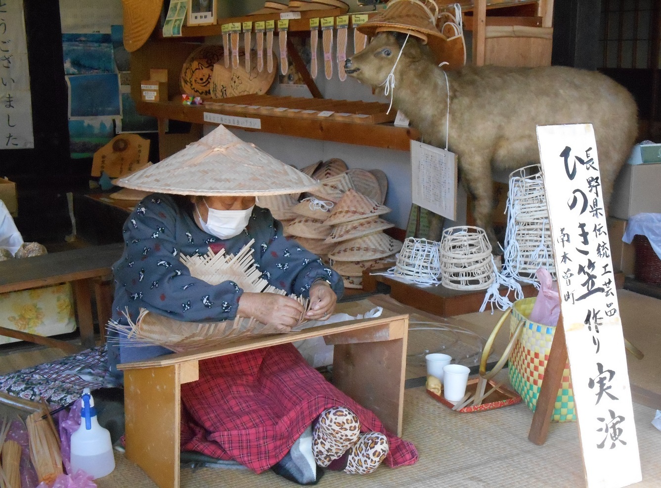 店先で伝統的な笠をかぶりマスクをした人物が竹細工を編んで作品が並んでいる
