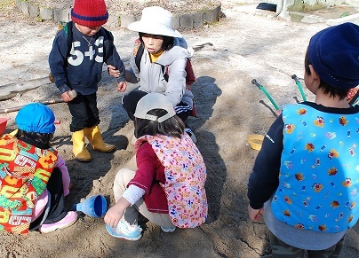 公園の地面で帽子をかぶった数人の幼児がリュックを背負いながら集まって遊んでいる様子