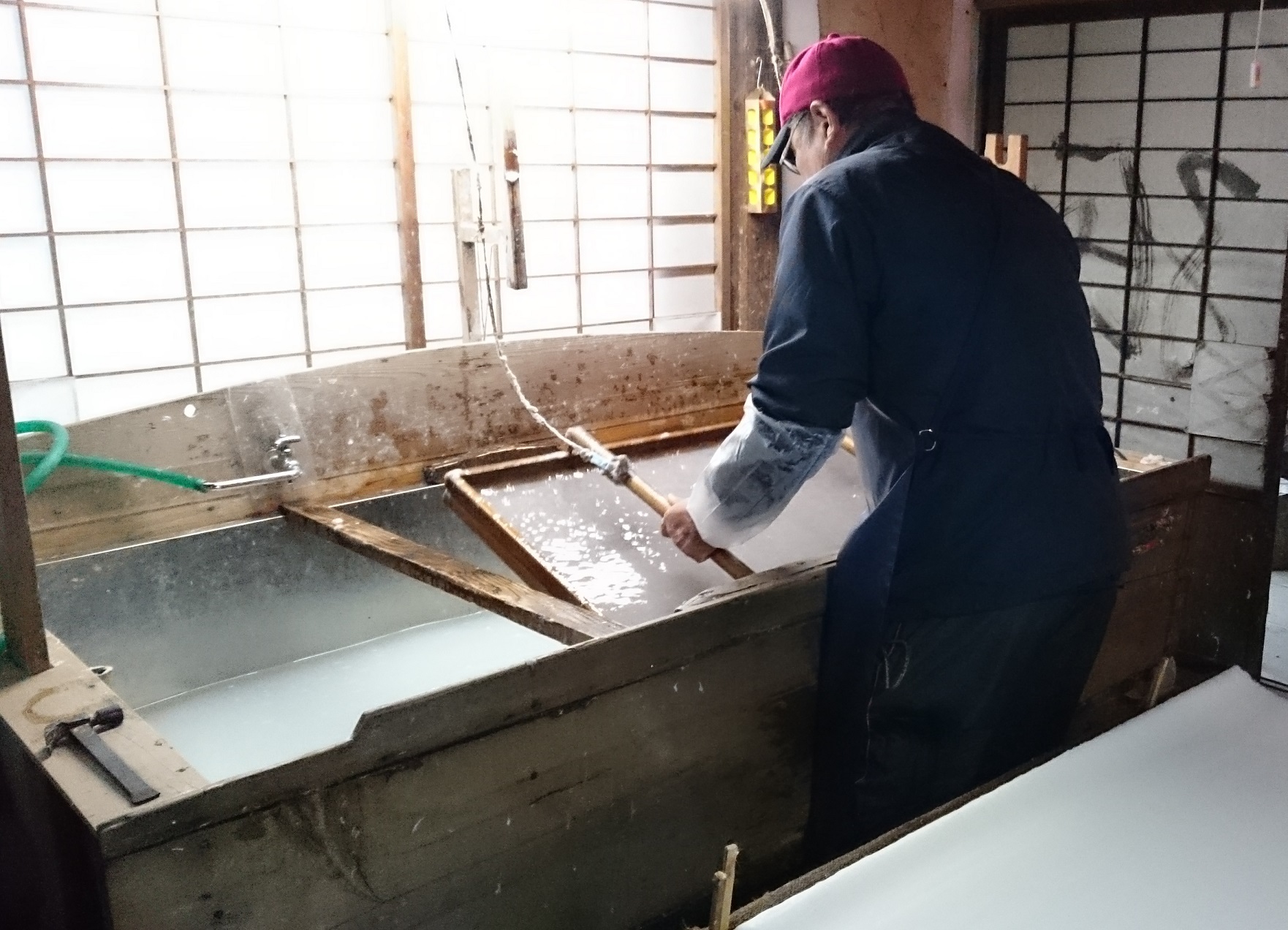 工場の作業場でキャップをかぶった作業員が大きな金属製の槽に向かってホースで水をかけながら洗浄している様子