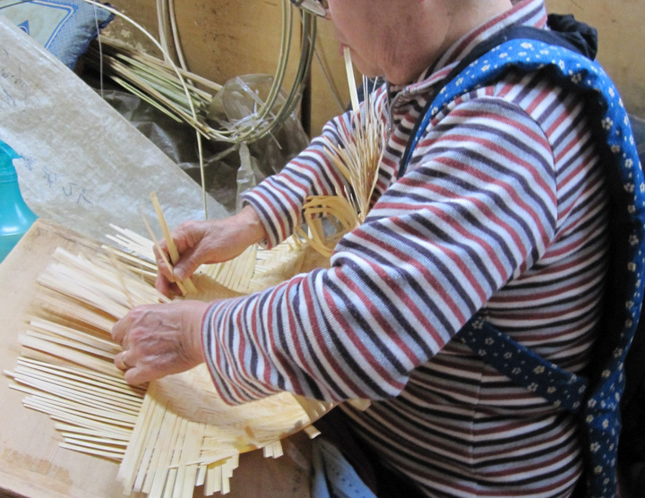 ストライプの長袖シャツを着た人が座って細長い紙や素材を折り編みしている様子