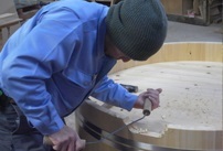 作業着の人物がニット帽をかぶり大きな木製の円形板を電動工具で削っている作業風景