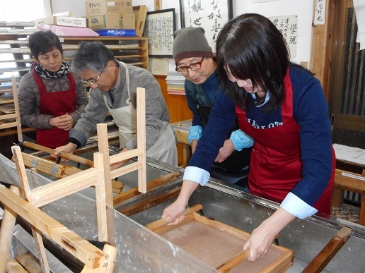 工房でエプロンを着た数人が版画の木枠に紙をのせて刷り作業を行っている様子