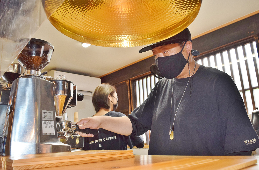 カフェのカウンターでマスクを着けた人物がエスプレッソマシンにポルタフィルターを取り付けている様子