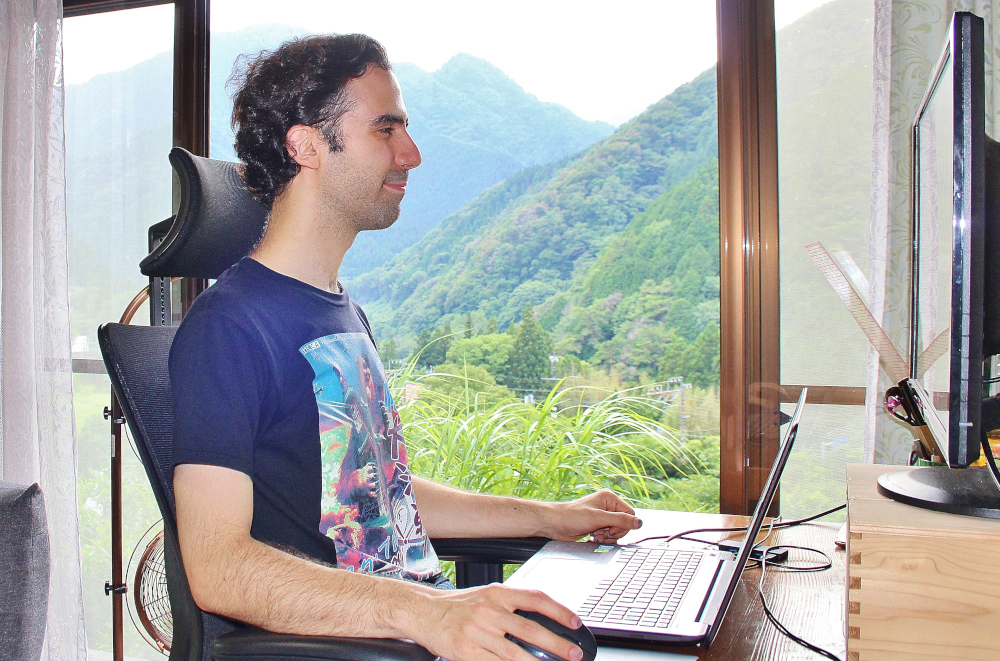 窓の外の山並みを背景にデスクでパソコンを操作する男性