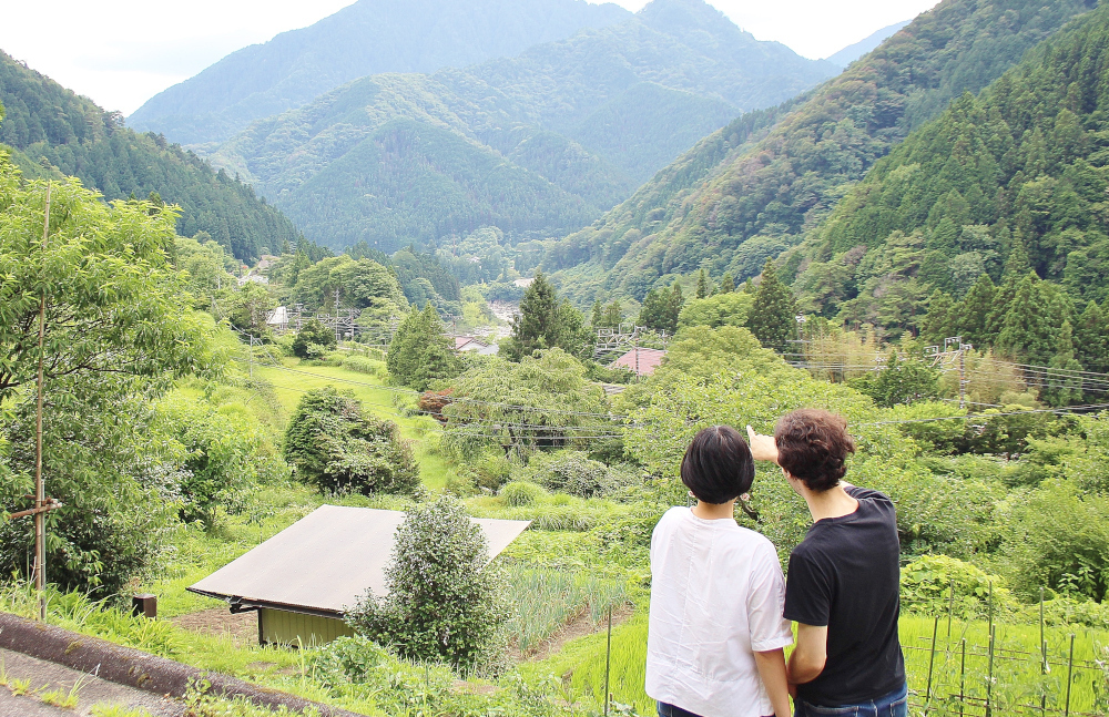 山あいの緑豊かな谷を背を向けた二人の人物が見つめる風景