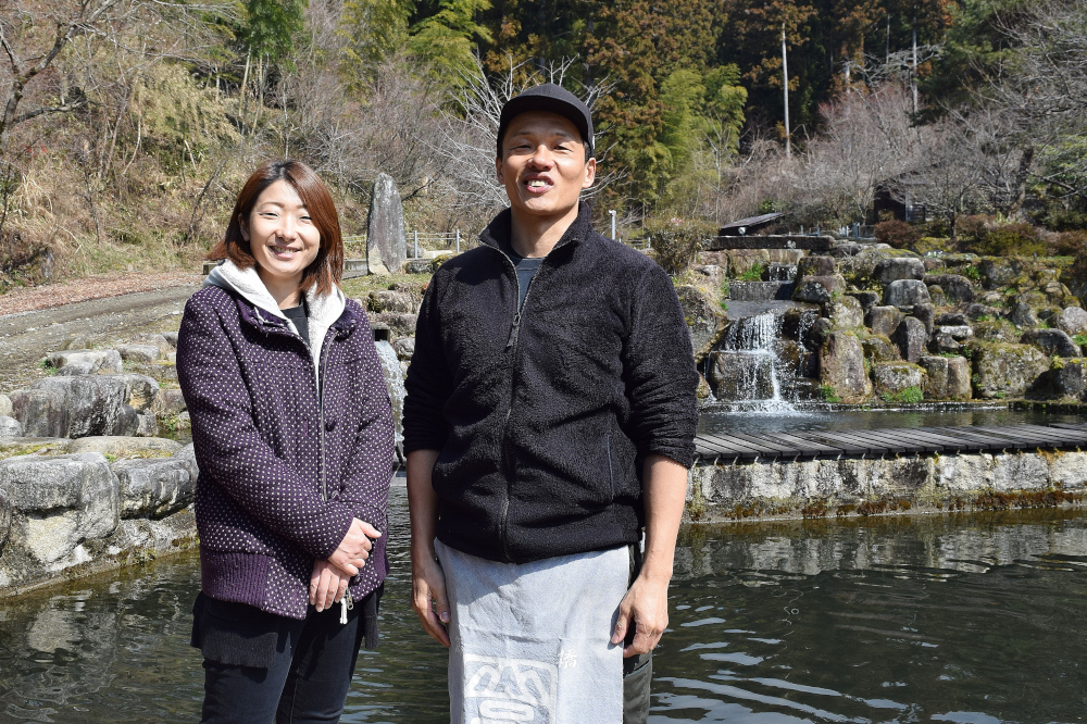 屋外の池の前でジャケットを着た二人の人物が並んで立っており背景に岩の滝と緑の木々が見える