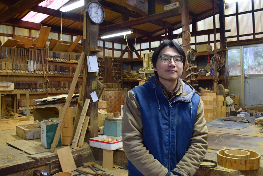 木工所の作業場で工具や木材が並ぶ作業台の前に立つ人物