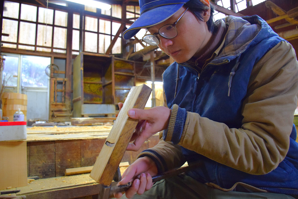 作業着の人物が工房の作業台で木材を手に持ち工具で削っている様子