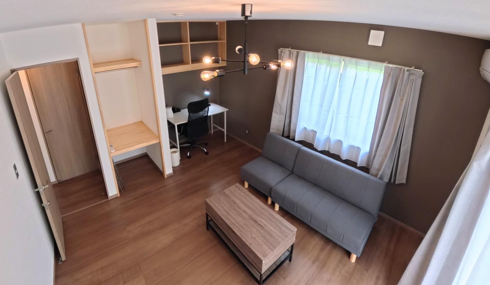 グレーの壁と大きな窓を備えた明るいリビングルームにグレーのソファと長方形のコーヒーテーブルが置かれ壁際にデスクと椅子と収納棚があり天井から複数の電球がぶら下がっている