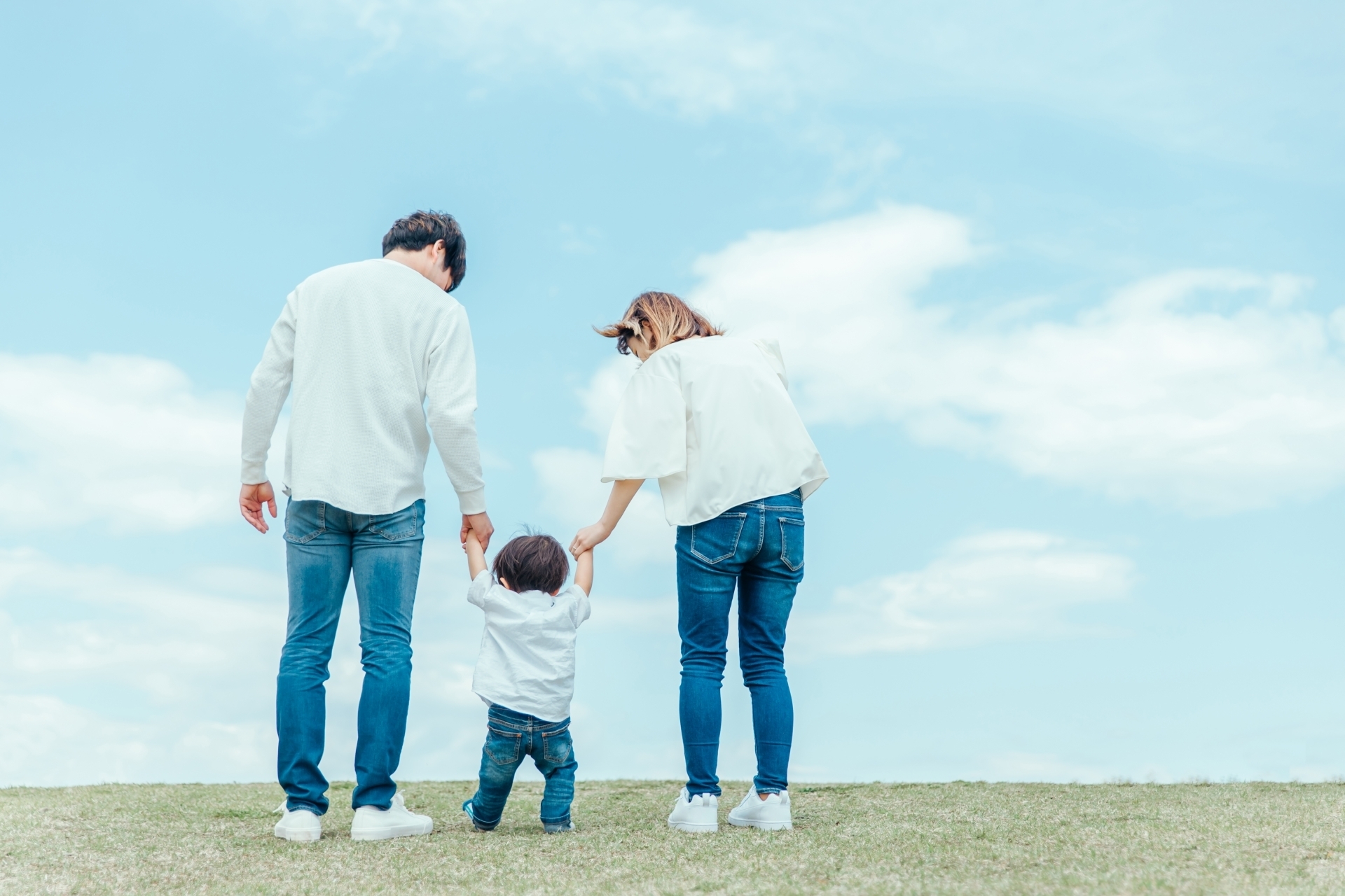 青空の下で白い服とジーンズを着た大人二人が両手を持って支えながら砂地を歩く幼児を後ろから見守る