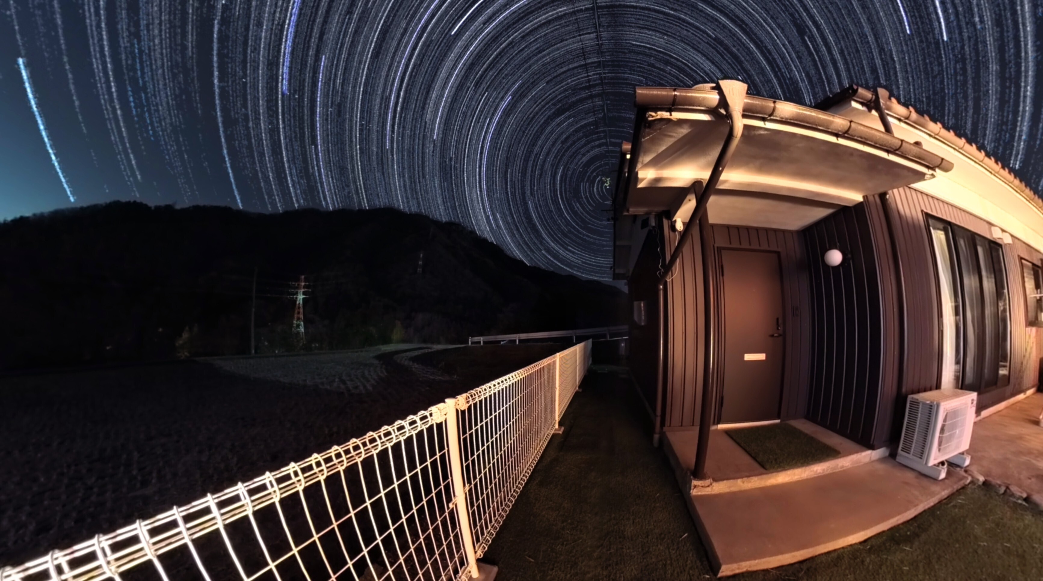 夜空に円を描く星の軌跡を背景に柵に沿った通路と軒先の明かりに照らされた小さな家の入口が並ぶ山間の夜景