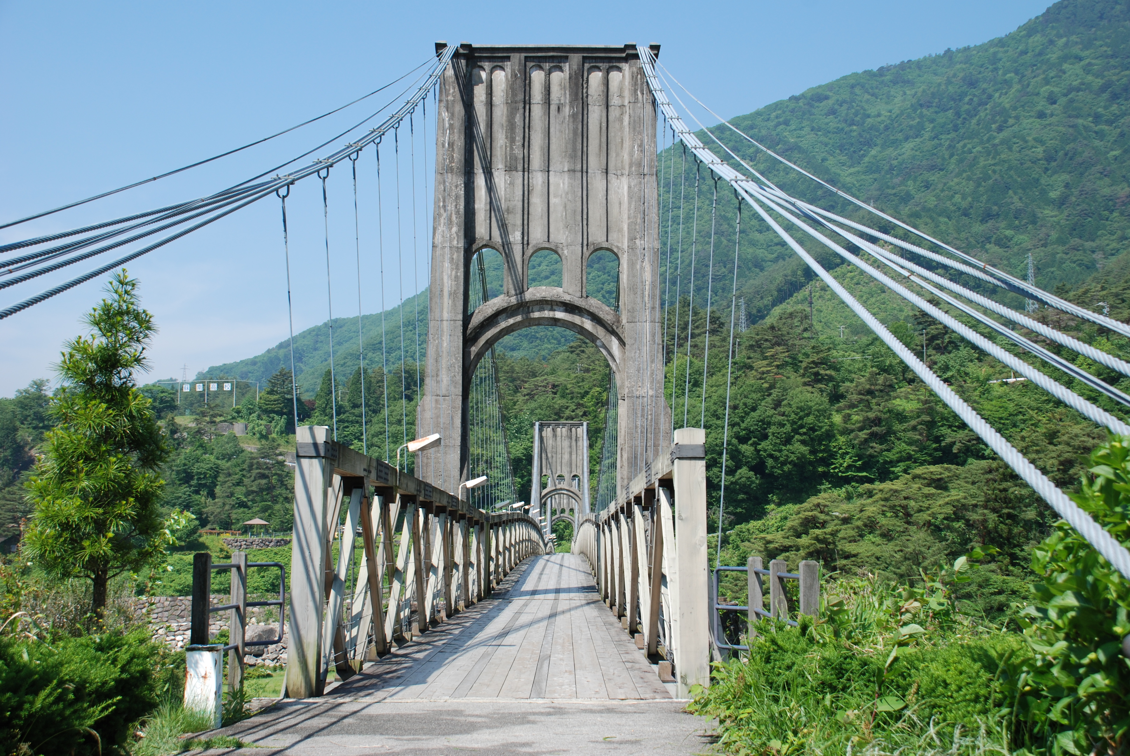 木製の歩道とワイヤーが張られたアーチ型の吊り橋が緑の山々に囲まれて続く風景