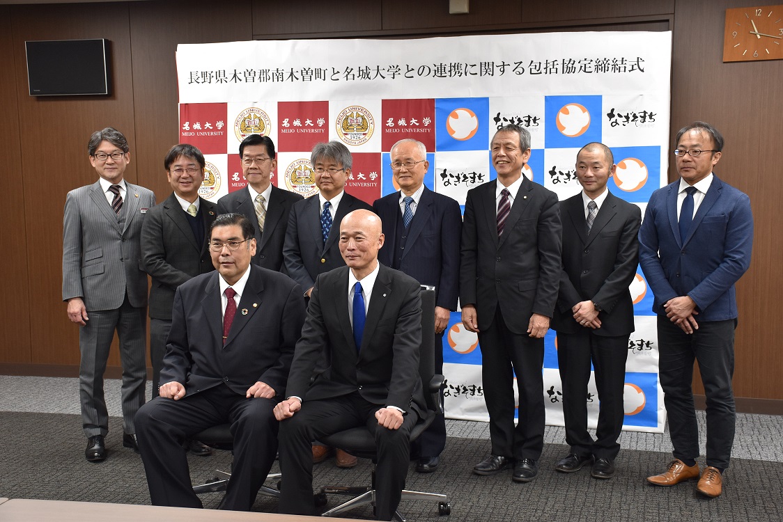 複数のビジネスマンがスーツを着て並んでいる記念写真で、背景にはロゴや文字がある。